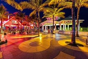A brightly lit outdoor area with palm trees, a stage with colorful lights, and a building with a sign reading "LAUDERHOLZBACH HOTEL". - Walking Distance to the Beach 3Br House With Pool (Lauderdale-by-the-Sea)