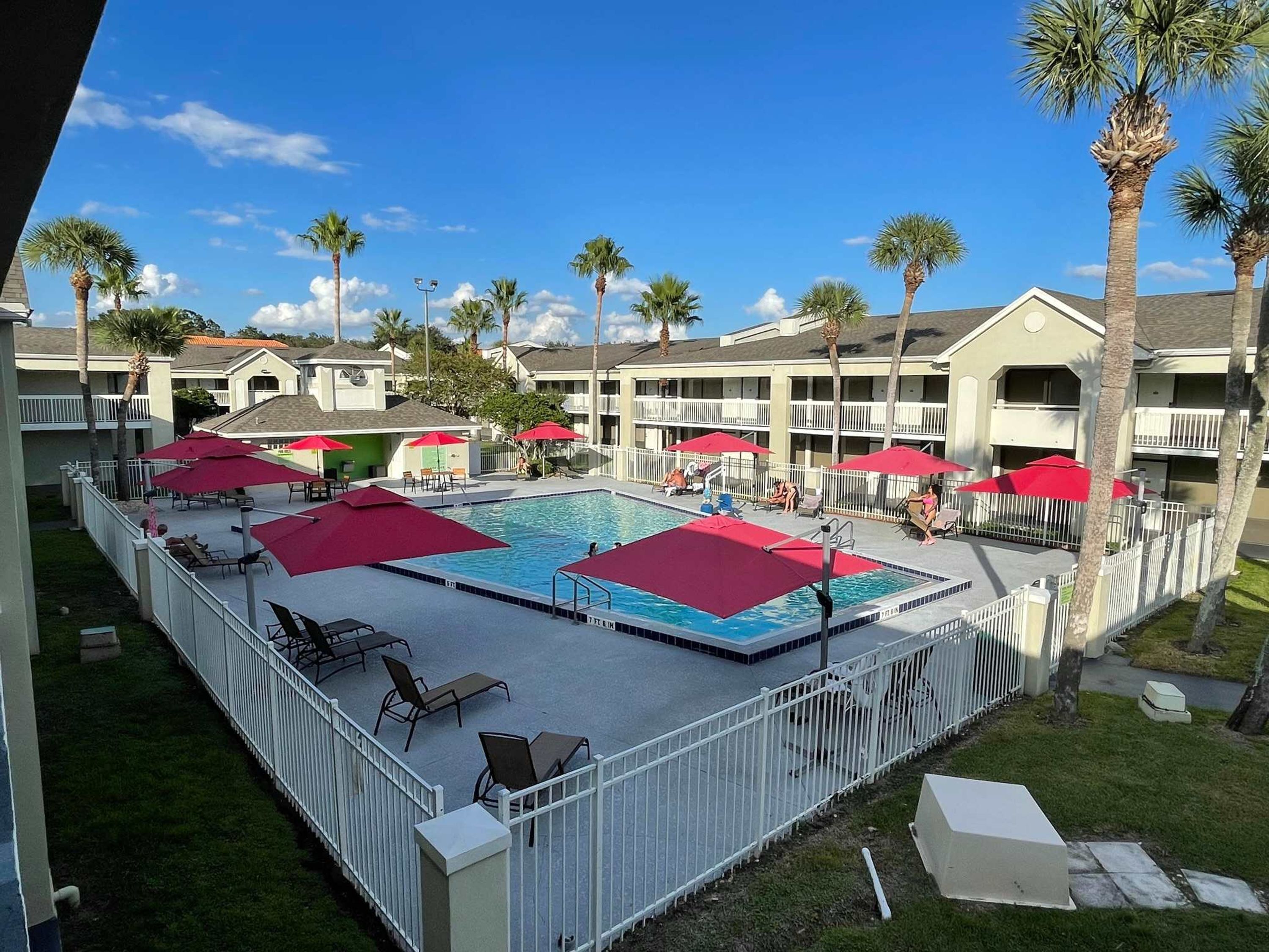 A swimming pool with red umbrellas, surrounded by a white fence, and palm trees.