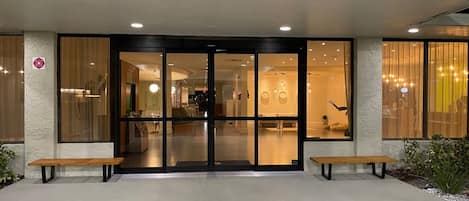 A hotel entrance with a glass door, a reception desk, and two benches outside.