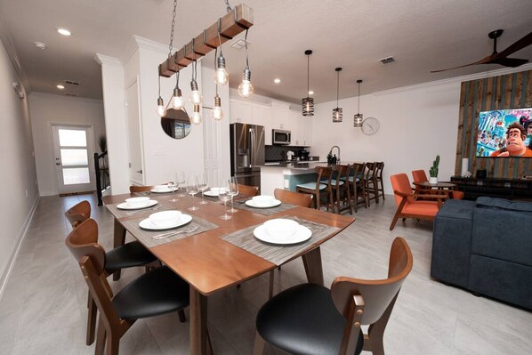 A modern dining area with a wooden table set for six, surrounded by chairs. A kitchen with a refrigerator and microwave is visible in the background.