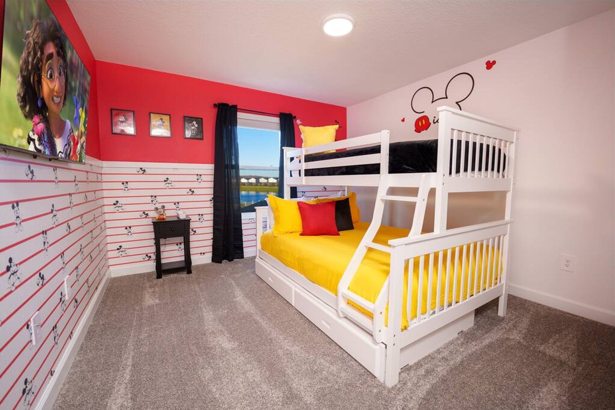 A bunk bed with yellow bedding, a small bedside table, a window with curtains, and a Mickey Mouse wall decal.