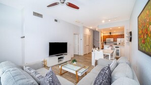 A modern living room with a sofa, coffee table, and TV. There is a kitchen in the background with cabinets and appliances. - Enchanted Townhome Near Disney Parks 3Br (Kissimmee)