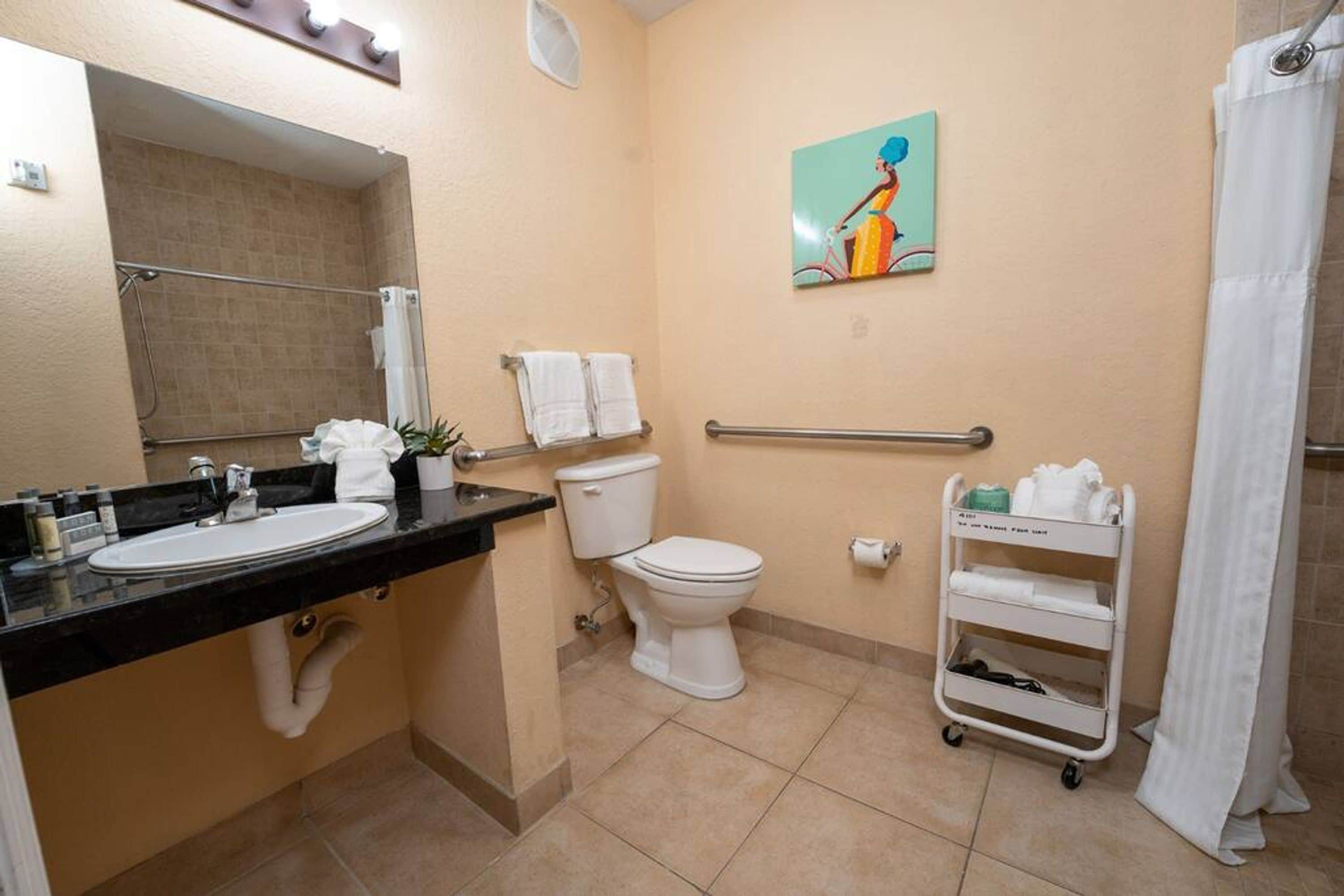Une salle de bain équipée d’un lavabo, de toilettes et d’une douche. On y trouve un porte-serviettes avec des serviettes, un tableau accroché au mur et un chariot contenant des articles de toilette.
