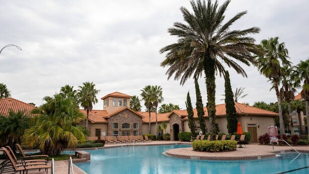 A swimming pool with lounge chairs, palm trees, and a building with a red-tiled roof. - Tuscana Resort 3 BR Near Disney (Davenport)