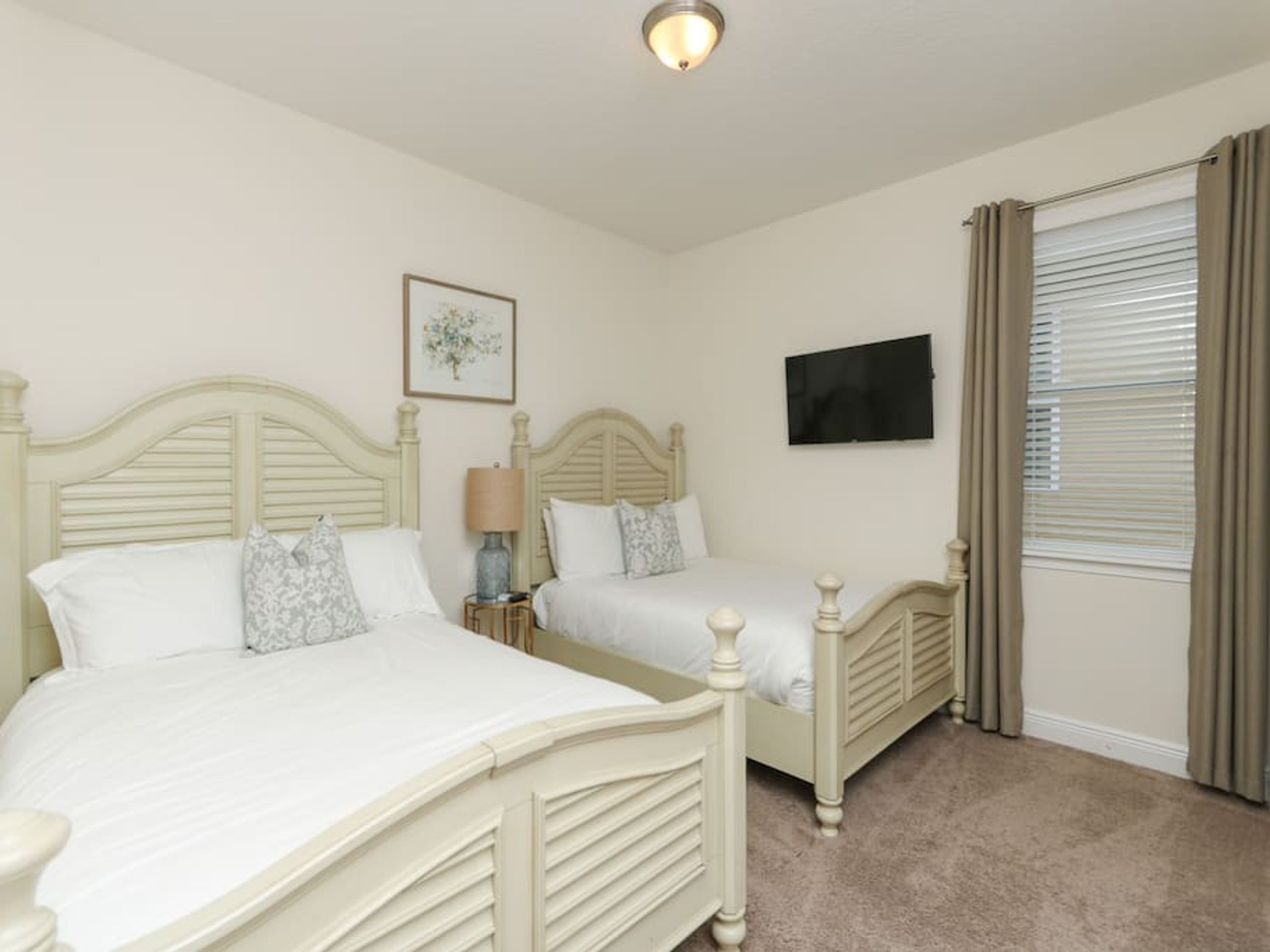 A bedroom with two beds, a bedside table with a lamp, a flat-screen TV, and a window with curtains.