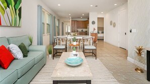Una sala moderna con sofá, mesa de comedor y sillas. Se ve una cocina al fondo con armarios y un refrigerador.