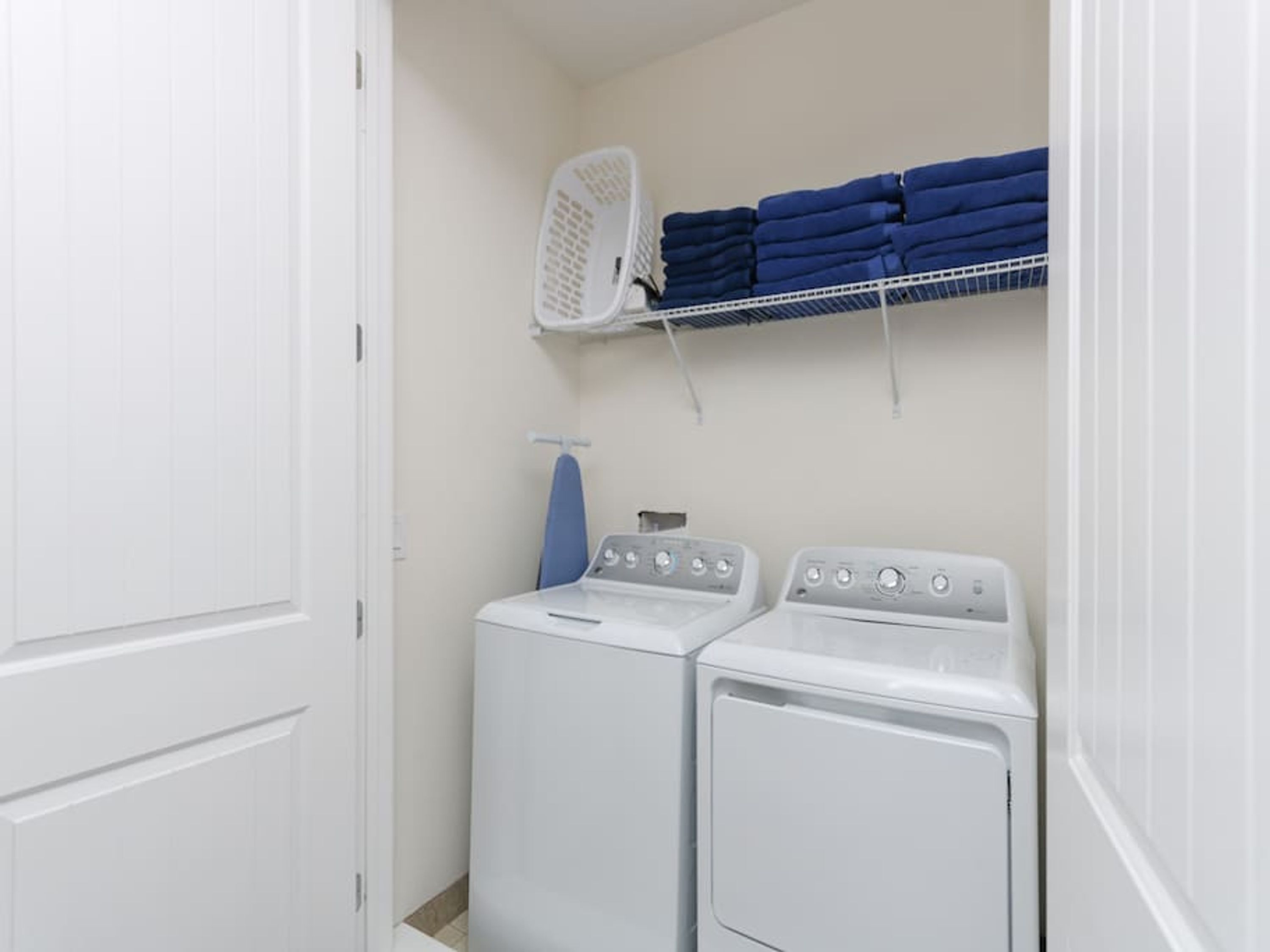 A laundry room with two white front-loading washers and dryers, a blue ironing board, and a white laundry basket on a shelf.