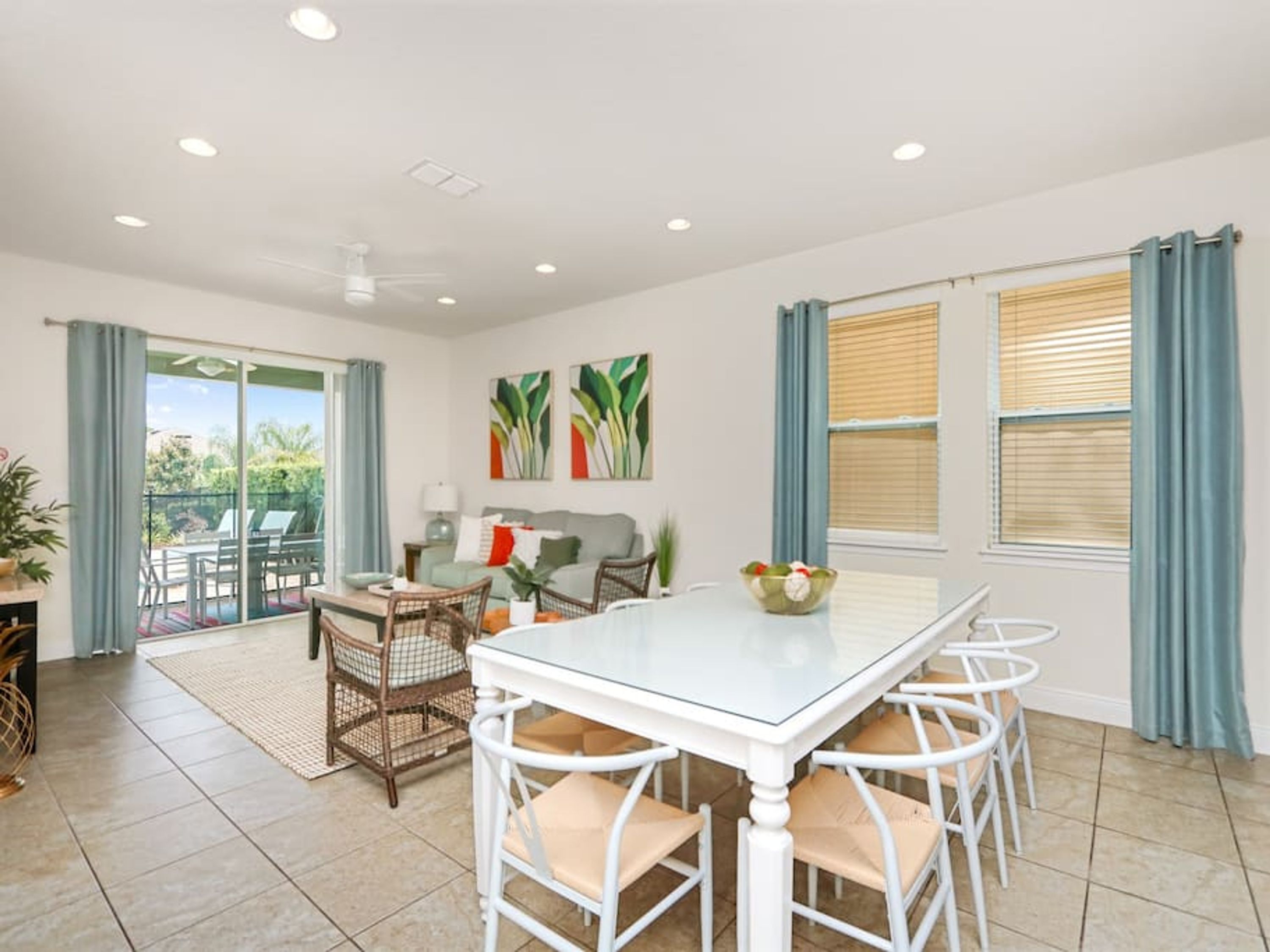 A spacious dining area with a glass-top table and white chairs, a sofa, and a balcony with a view.
