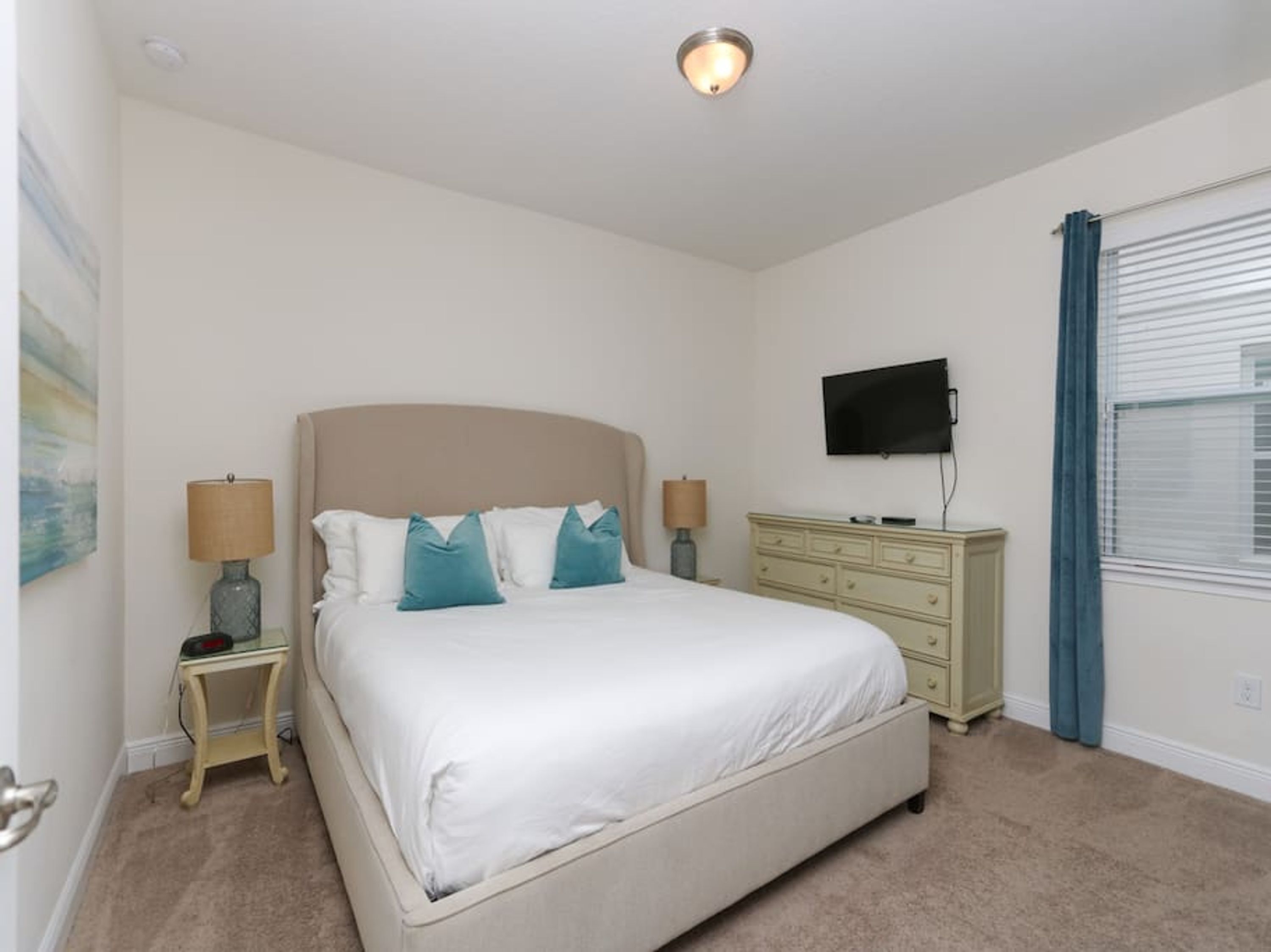 A bedroom with a bed, two bedside tables, a dresser, a television, and a window with blinds.
