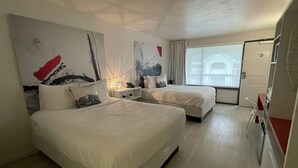 Ett hotellrum med två sängar, ett litet skrivbord, en stol och ett fönster med utsikt över en byggnad.