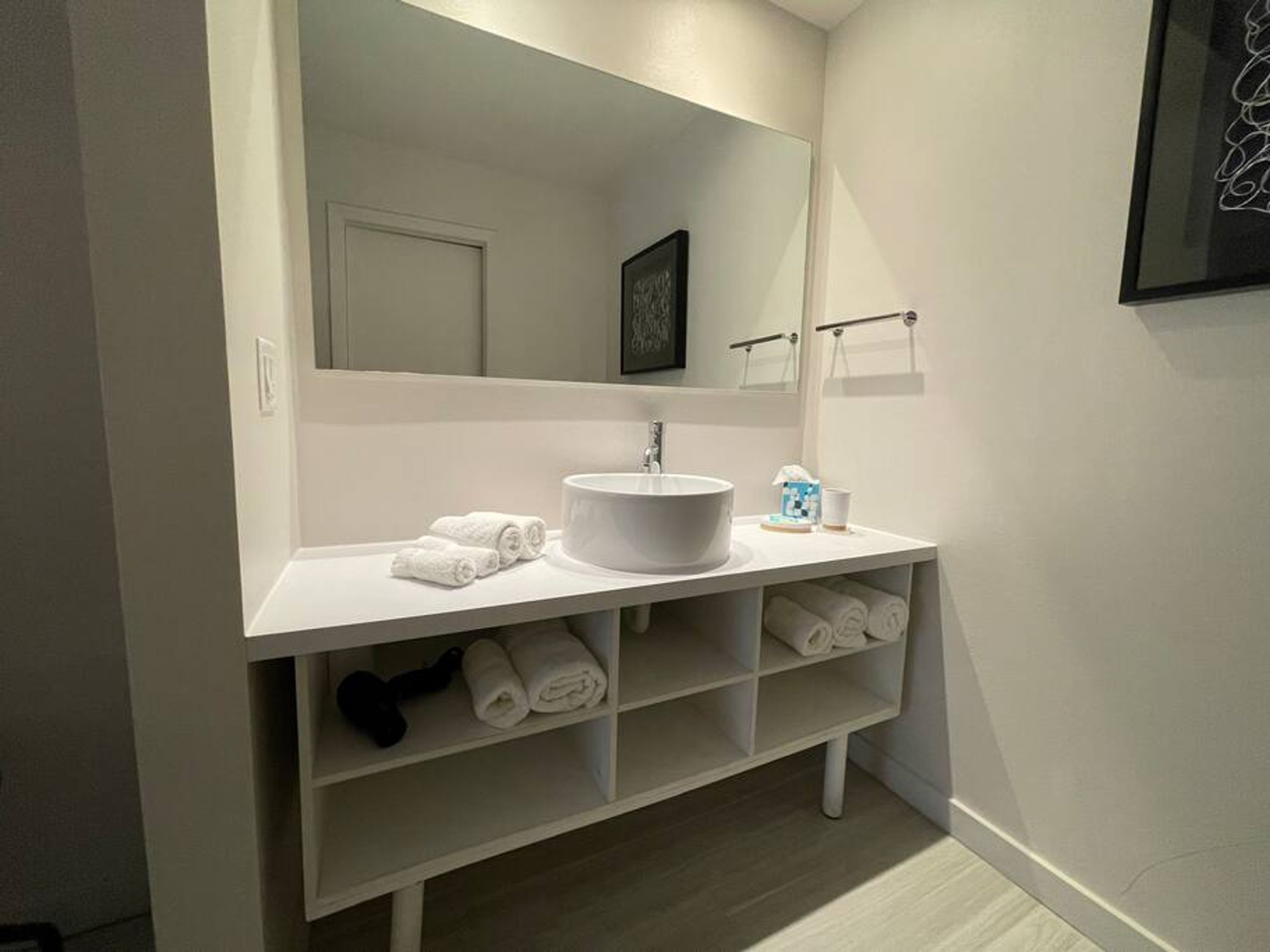 A modern bathroom with a white vanity, a round sink, and a large mirror. There are towels and toiletries on the counter.