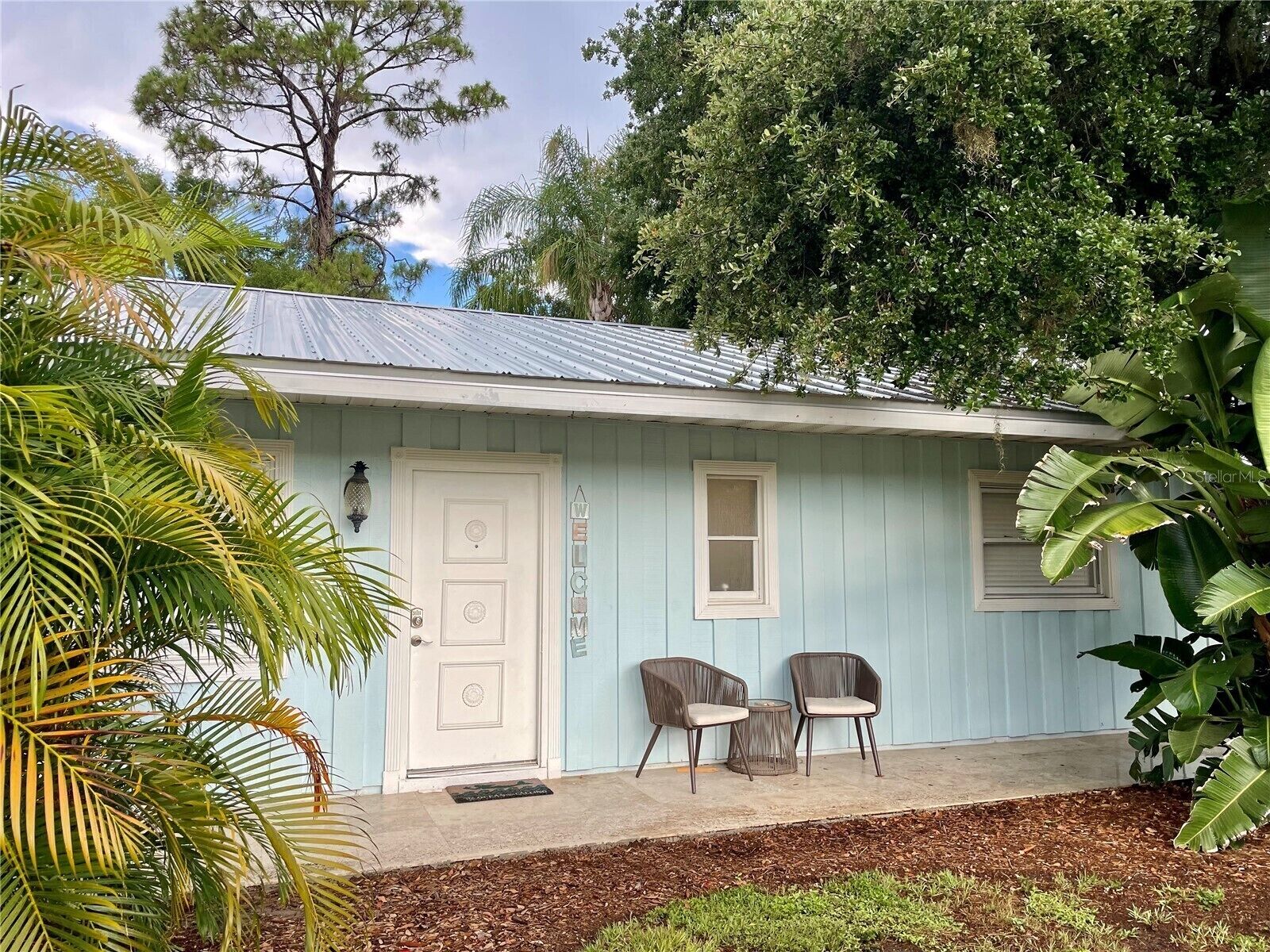 A light blue house with a white door, two windows, and a porch with two chairs.