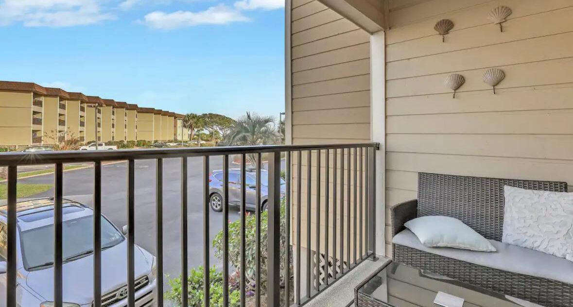 A balcony with a wicker sofa and a view of a parking lot and hotel buildings.