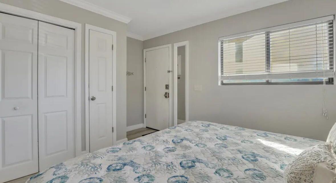 Un dormitorio con cama, dos puertas, una ventana con persianas y un interruptor de luz en la pared.