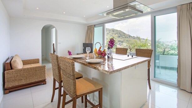 Una cocina moderna con una isla central, zona de comedor con muebles de mimbre y ventanas amplias con vistas a un paisaje montañoso.