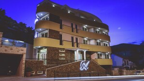 A multi-story hotel with a prominent illuminated logo on the facade, a well-lit entrance, and a parking area in front. - Hotel Atina by ENTOPYA (Budva)