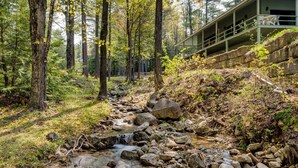 Un área boscosa con un arroyo, rocas y un edificio con un balcón que da al arroyo.