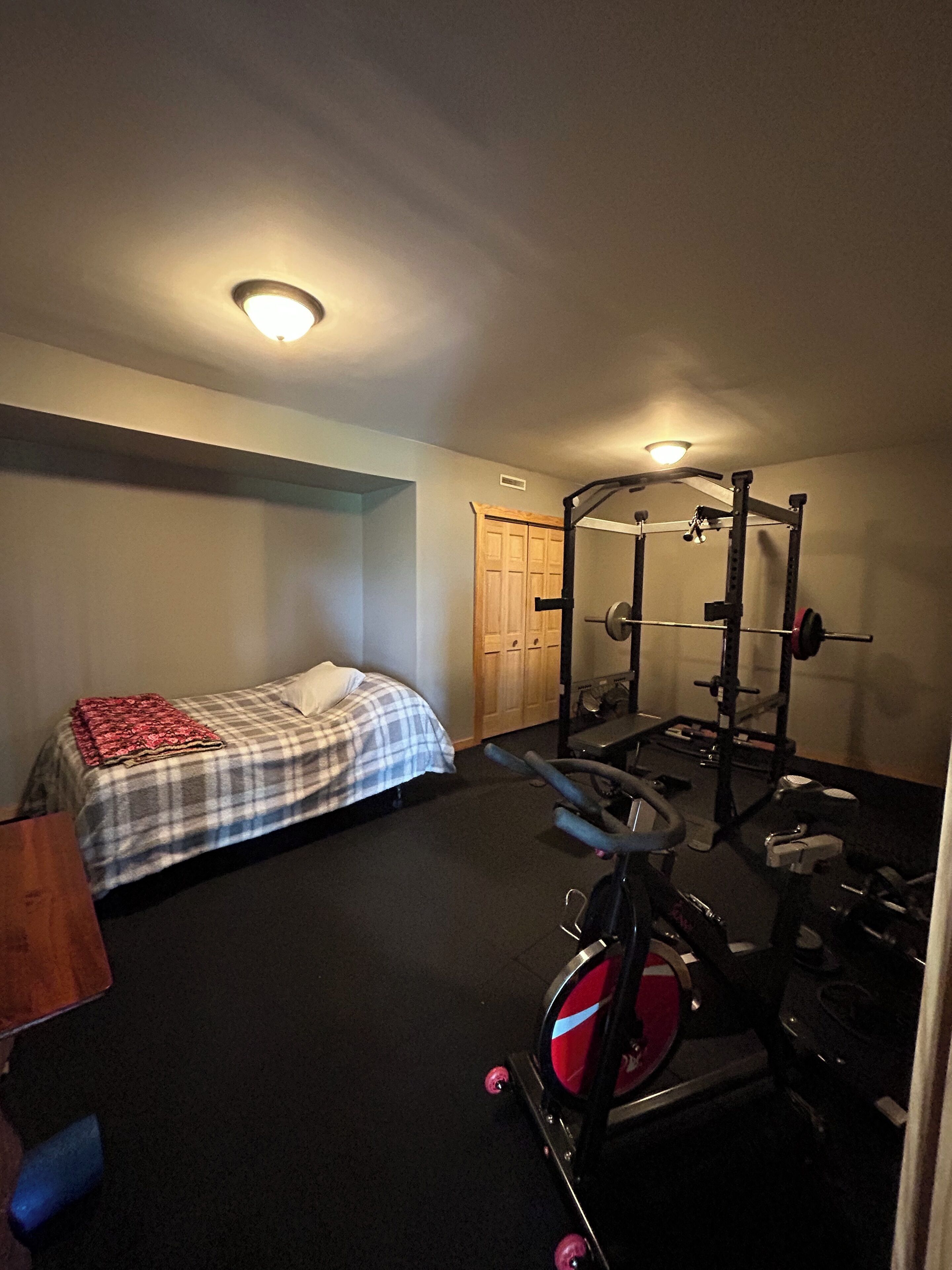 Un dormitorio con cama, un espacio para ejercitarse con una prensa de pesas y una bicicleta estática, y una puerta que da a otra habitación.