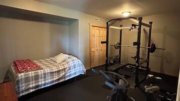 Un dormitorio con cama, un espacio para ejercitarse con una prensa de pesas y una bicicleta estática, y una puerta que da a otra habitación.