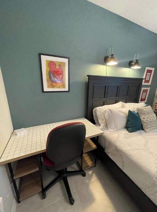 A bedroom with a bed, a desk, and a chair. There is a framed picture on the wall and two wall-mounted lamps.