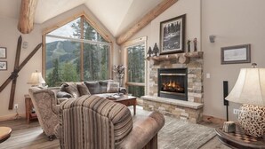 A cozy living room with a stone fireplace, large windows, and a high ceiling with exposed beams. - Lakota Lookout | 4BR Hot Tub, Across from Resort (Winter Park)