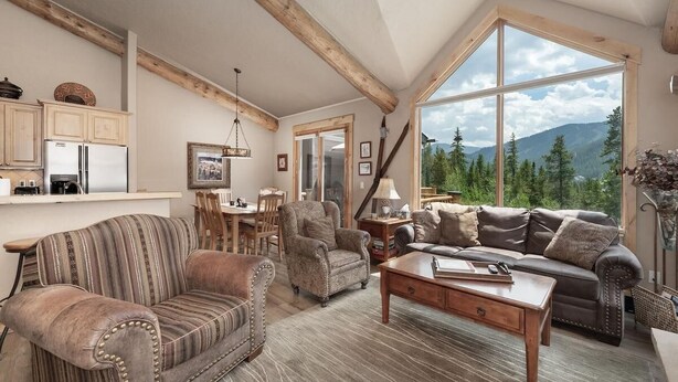 A cozy living room with a large window, wooden beams, a sofa, a coffee table, and a dining area. - Lakota Lookout | 4BR Hot Tub, Across from Resort (Winter Park)