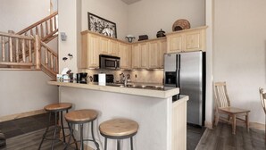 A kitchen with wooden cabinets, a stainless steel refrigerator, a microwave, and two bar stools. - Lakota Lookout | 4BR Hot Tub, Across from Resort (Winter Park)
