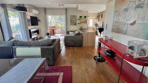 A modern living room with a sofa, a dining table, and a kitchen area in the background. - Artistic 2-bedroom apartment in green & peaceful Vari (Vari)