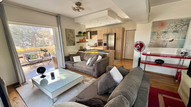 A modern living room with a sofa, coffee table, and a dining area with a red table and chairs. - Artistic 2-bedroom apartment in green & peaceful Vari (Vari)