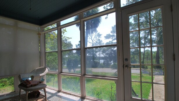 Ein Zimmer mit großen Fenstern, einem Korbstuhl und Blick auf eine Wiese und Bäume.