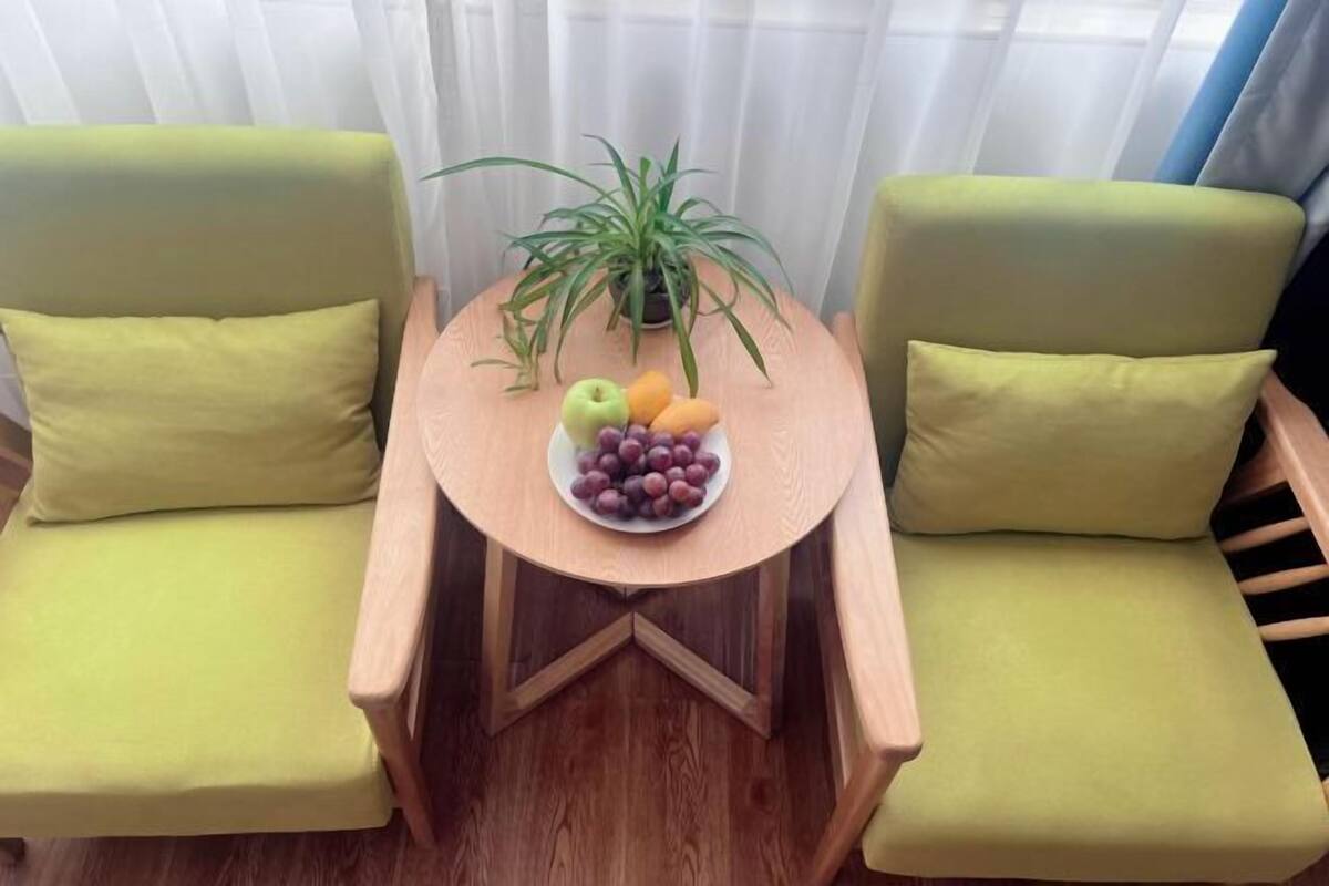 A room with two green chairs, a small round table with a fruit bowl, and a potted plant.