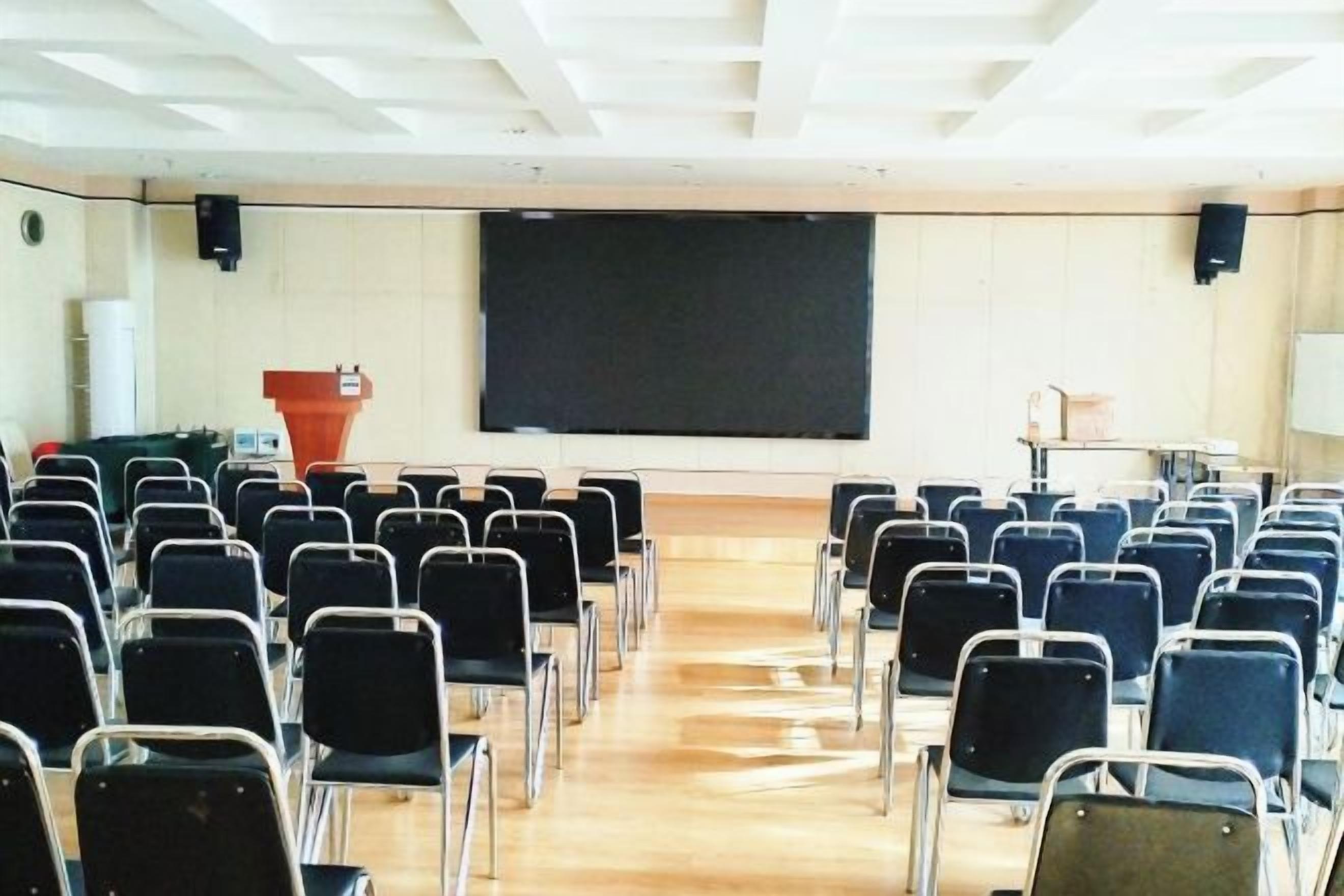 Un salón de conferencias con filas de sillas mirando una pizarra, un atril y una pantalla de proyector.
