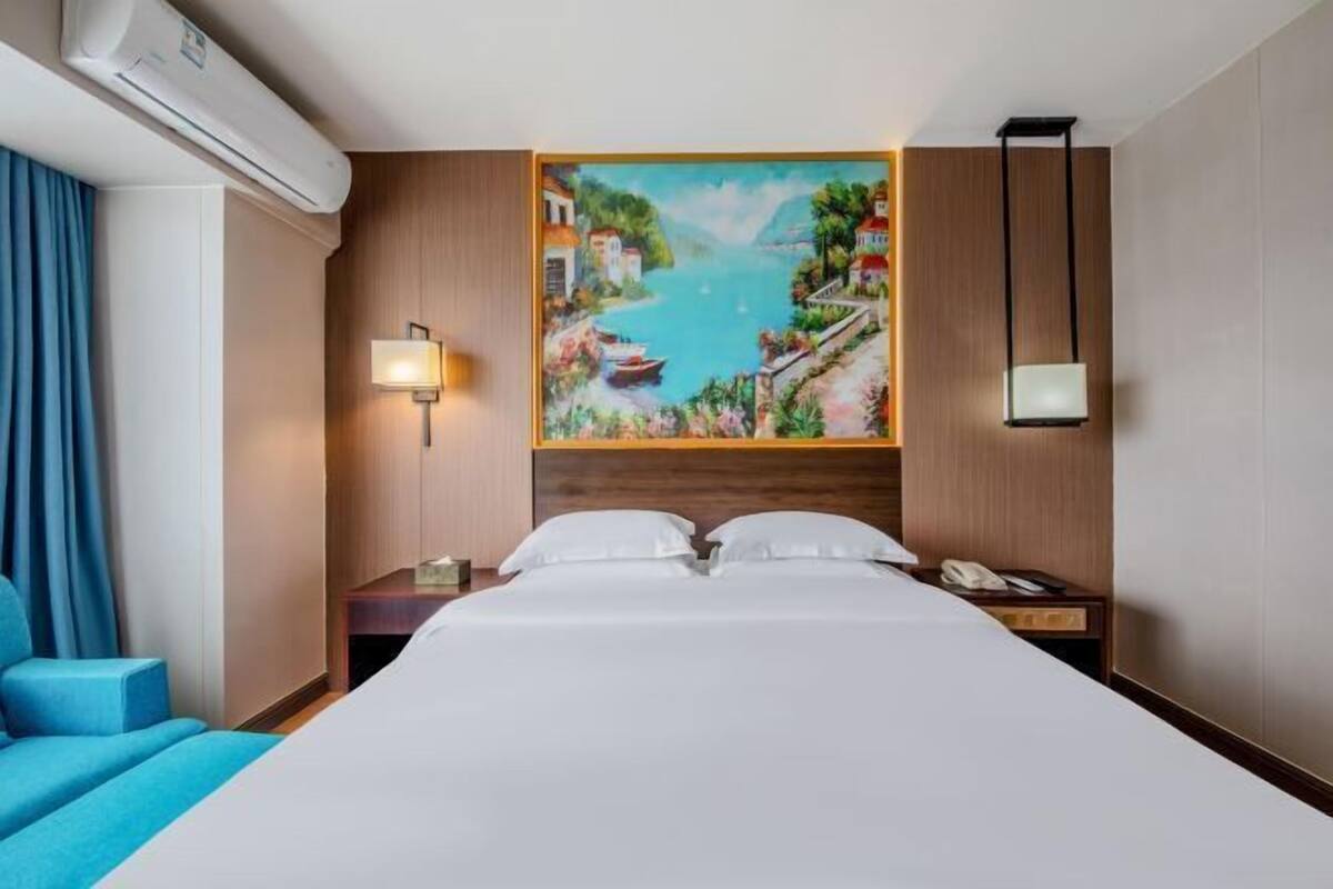 A hotel room with a large bed, a painting of a coastal scene, a bedside lamp, and a nightstand with a phone.