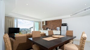 Ein moderner Essbereich mit einem dunklen Holztisch und Korbstühlen, eine Küche im Hintergrund und ein Blick auf die Stadt durch große Fenster.