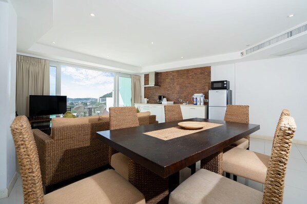 A modern dining area with a dark wooden table and wicker chairs, a kitchen in the background, and a city view through large windows.