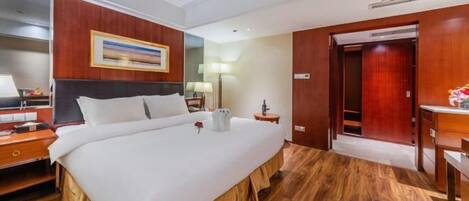 A hotel room with a large bed, wooden flooring, a painting on the wall, and a desk with a lamp.