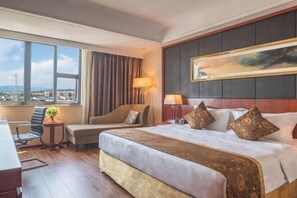 A hotel room with a large bed, a desk, a chair, a view of the city, and a painting on the wall.
