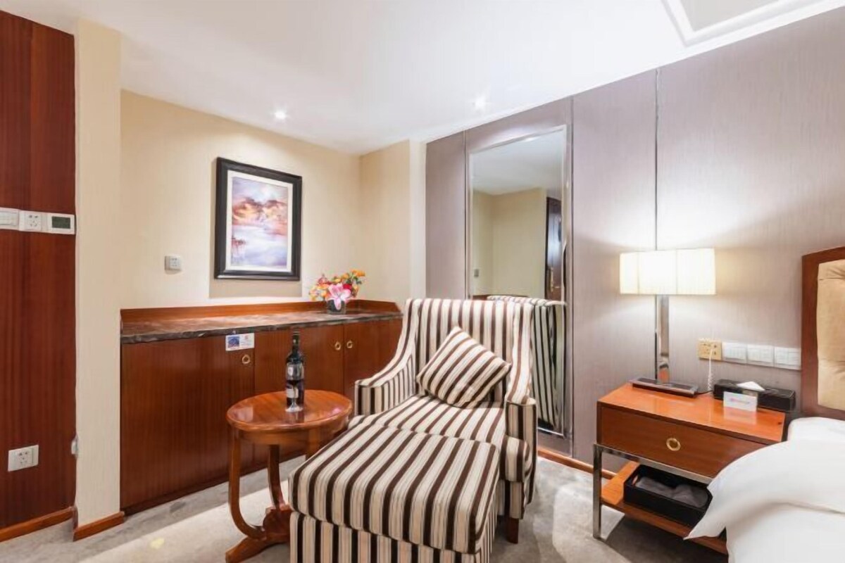 A hotel room with a bed, a bedside table, a chair, a desk, a mirror, and a wall-mounted artwork.