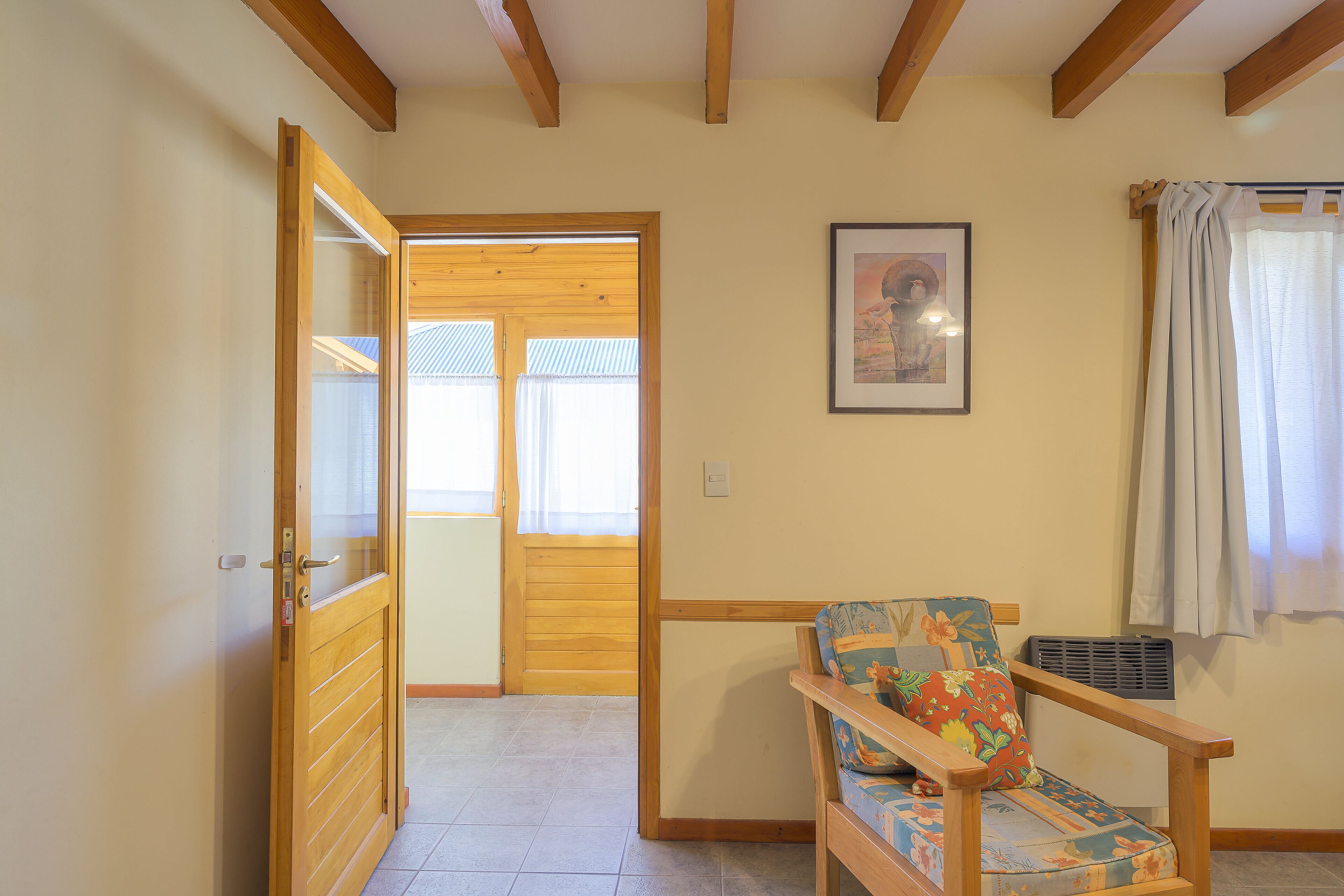 A room with a wooden door, a chair with a floral cushion, a framed picture on the wall, and a window with curtains.