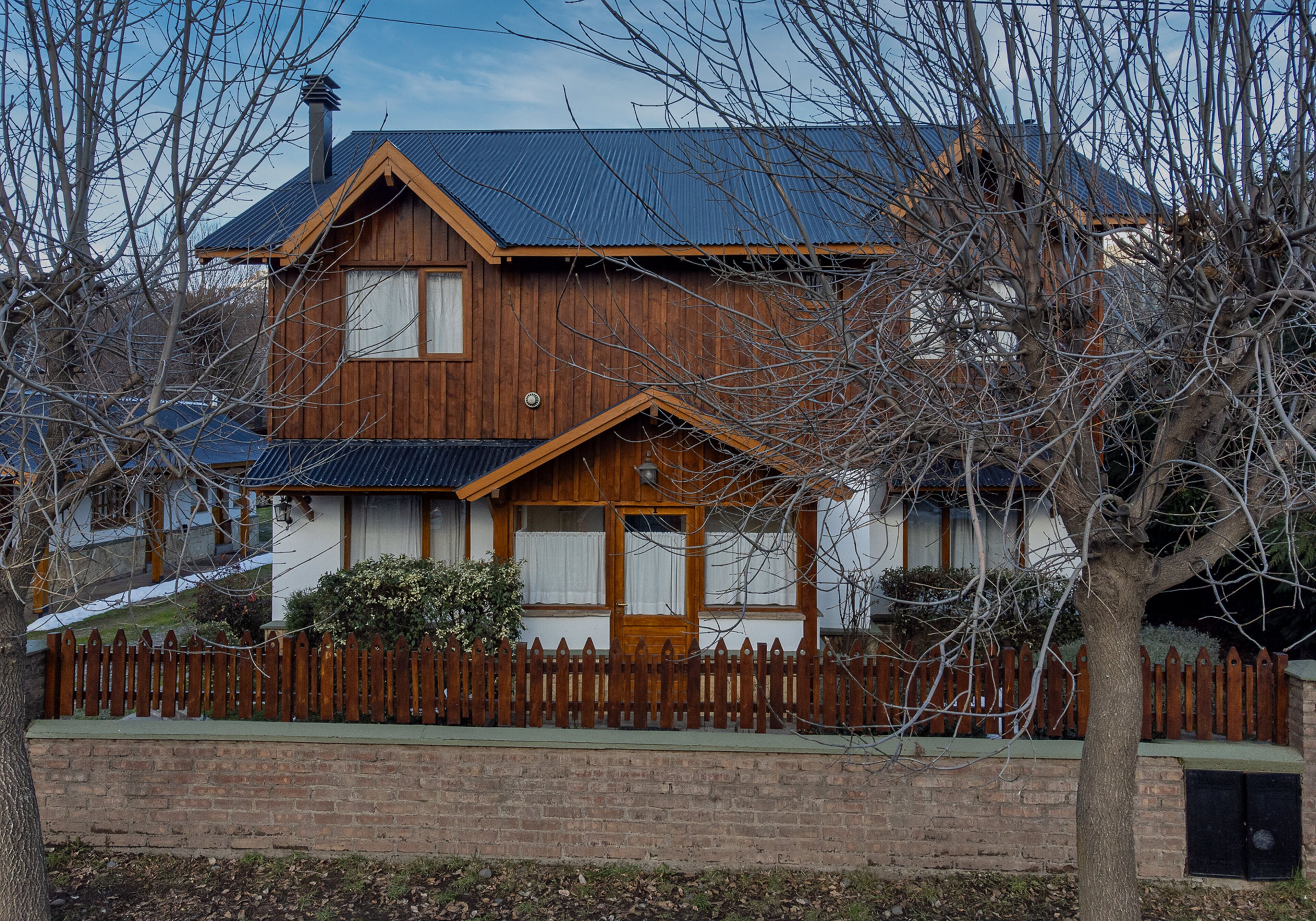 Une maison en bois à deux étages, avec un toit à pignon, des fondations en briques et une clôture en bois à piquets.