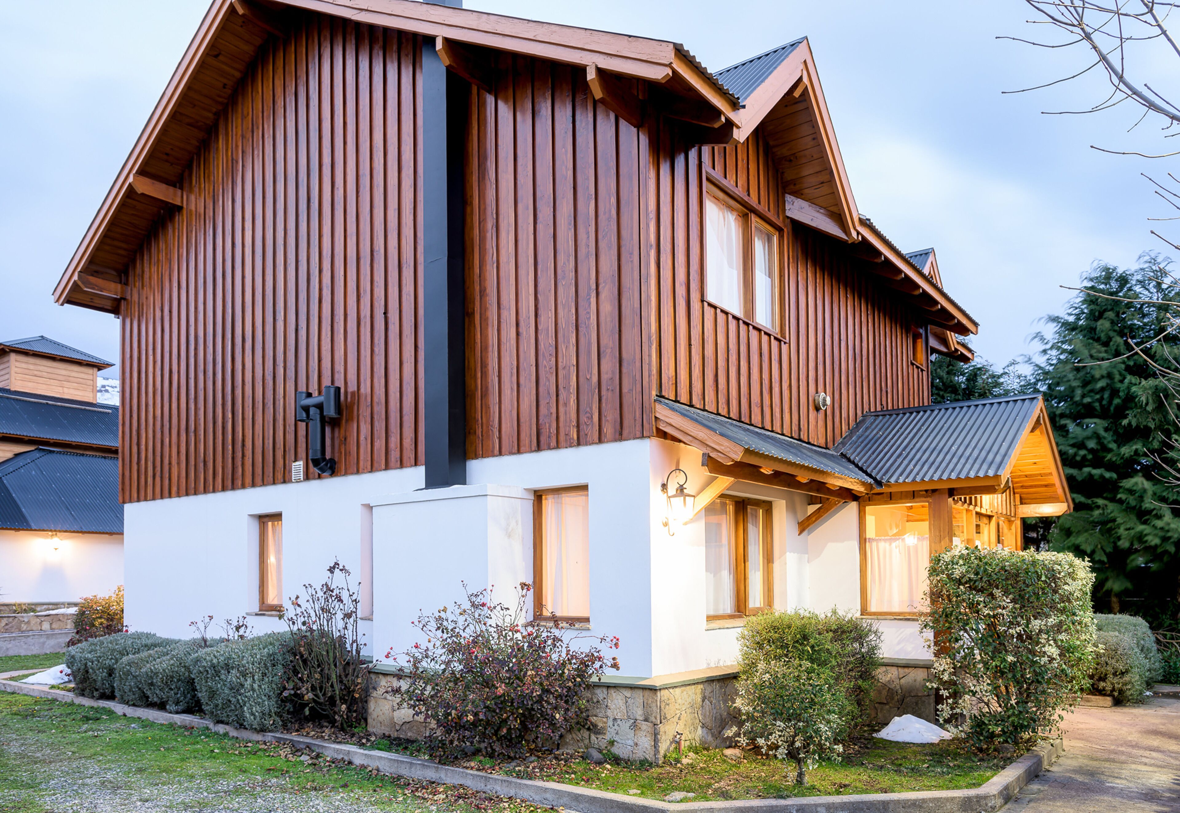 A two-story wooden house with a white facade, a chimney, and a well-maintained garden with hedges and shrubs.