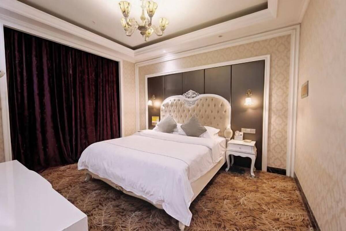 Een slaapkamer met een groot bed, een nachtkastje, een kroonluchter en een kamerbreed tapijt met een patroon.
