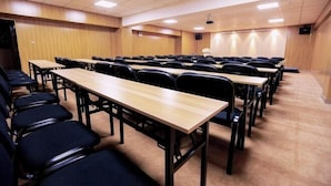 Una sala de conferencias con filas de mesas y sillas, equipada con un proyector y una pantalla.