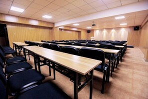 Une salle de conférence avec des rangées de tables et de chaises, équipée d’un projecteur et d’un écran.