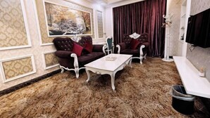 Una habitación con dos sillones, una mesa de centro blanca, un cuadro grande, una cortina roja y una araña.