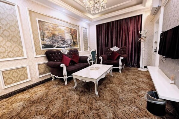Een kamer met twee fauteuils, een witte bijzettafel, een groot schilderij, een rood gordijn en een kroonluchter.