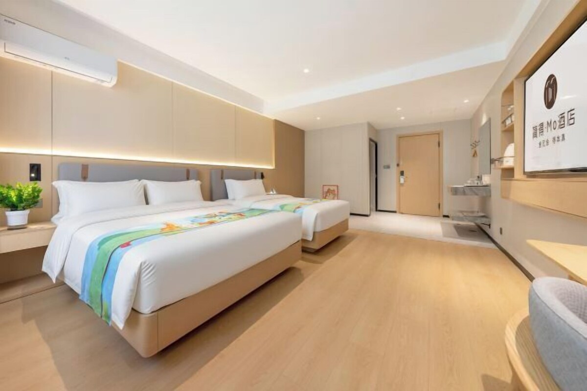 Una habitación de hotel moderna con dos camas, una mesita de noche, una planta en maceta y un televisor montado en la pared.