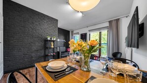 A dining area with a brick wall, a wooden table set for two, a vase with flowers, and a television mounted on the wall. - Warsaw Leszno Apartment with Parking - by Rentujemy (Warszawa)