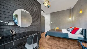 A modern bedroom with a brick wall, a round mirror, a bed with colorful pillows, and a desk with a chair. - Warsaw Leszno Apartment with Parking - by Rentujemy (Warszawa)