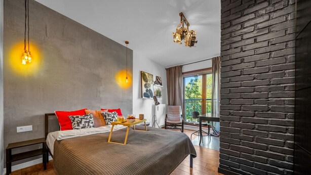 A modern bedroom with a large bed, a wooden bedside table, a bench, a hanging light fixture, a black brick accent wall, and a view of the outdoors. - Warsaw Leszno Apartment with Parking - by Rentujemy (Warszawa)